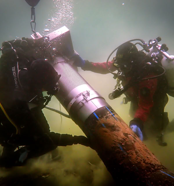 Deux plongeurs en tenue de plongée travaillent sous l'eau sur un grand tuyau métallique.