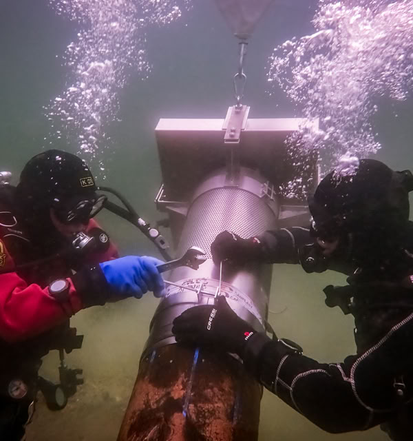 Deux plongeurs en combinaison de plongée travaillent sous l'eau sur une grande structure métallique, avec des bulles d'air s'élevant autour d'eux.
